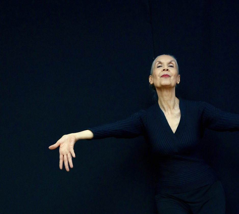 A woman wearing black extends her arm with her hand and palm extended out and down with fingers open. She is against a black backdrop.