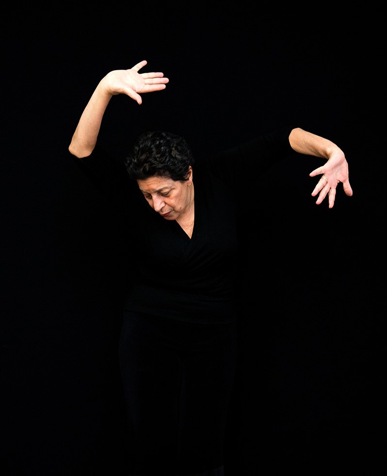 A woman wearing black with arms and hands open and angled around her head dramatically. She is against a black backdrop.