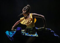 A dancer in a yellow shirt and blue skirt whips her braids to the side under dramatic lighting.