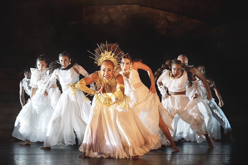 Several dancers wearing white and in a triangle formation lunge forward.