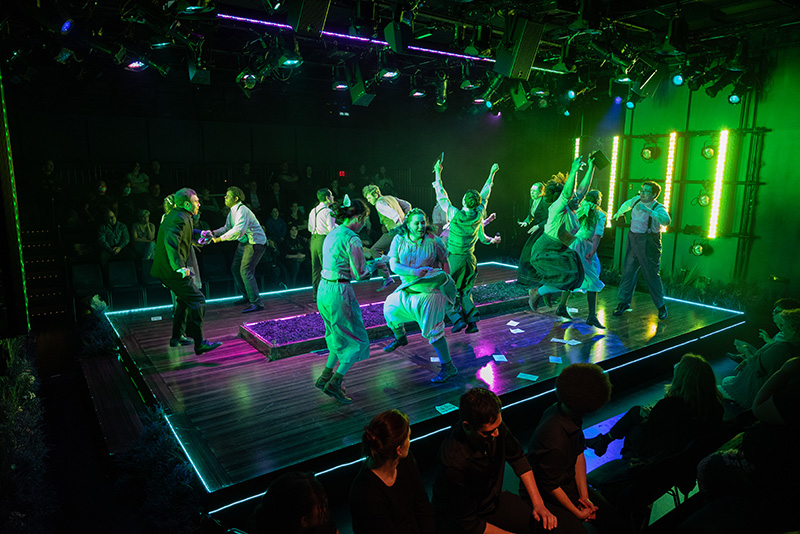 A group of performers jump around a stage in green light. The stage has audience members in the round.