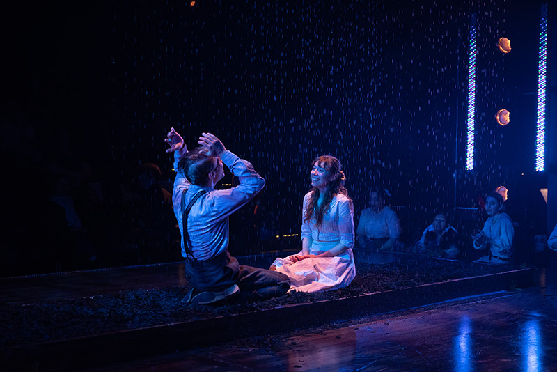 Two performers sit onstage facing each other under blue light that appears to be raining. The stage is in the round.