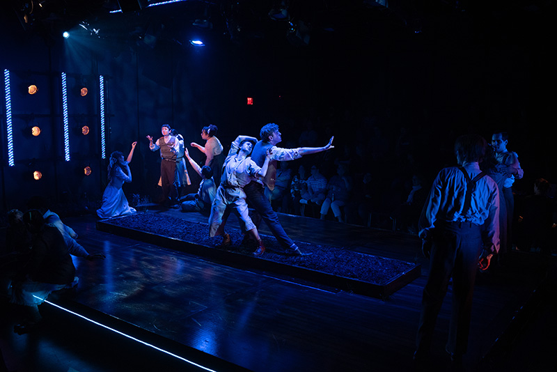 A handful of performers pose dramatically under harsh blue light on a stage in the round,