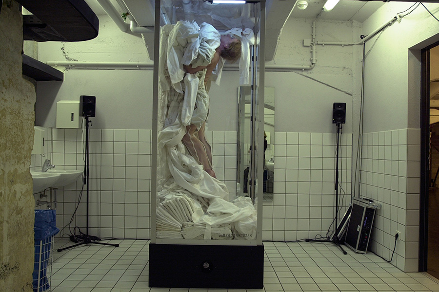 One dancers moves slowly with fabric in a vertical glass rectangle in a public bathroom.