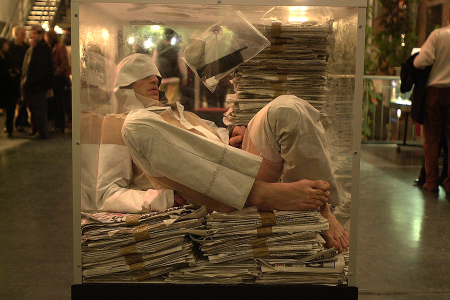 A dancer in a glass cube filled with newspaper is scrunched down against the glass. A busy metropolitan scene is behind him. 