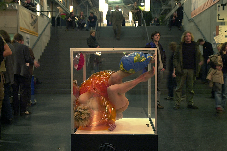 A woman in a glass cube moves almost upside down. Around her, people walk up and down public stairs.