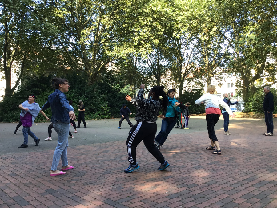 About ten dancers stand and move on brick pavement in the shade of trees.