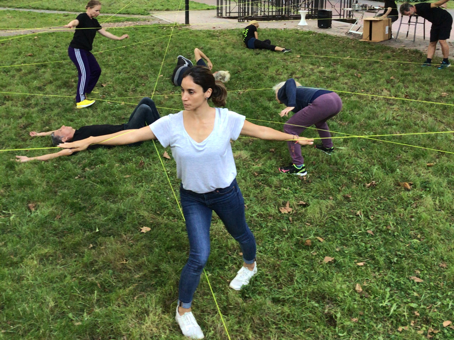 Seven dancers on grass are moving around and amongst a series of taut strings.