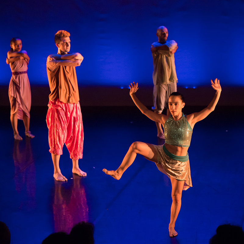 One dancer stands on one leg with her other extended and bent, both arms up and out. Three other dancers stand facing her with crossed arms on a blue lit stage.