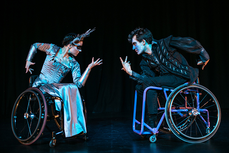 Two dancers in wheelchairs face off on a black stage. One is wearing a silver dress and the other is wearing black.