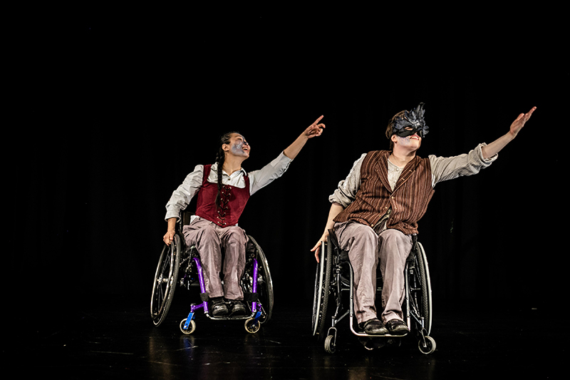 Two dancers in wheelchairs on a black stage lean to one side with one arm extended.