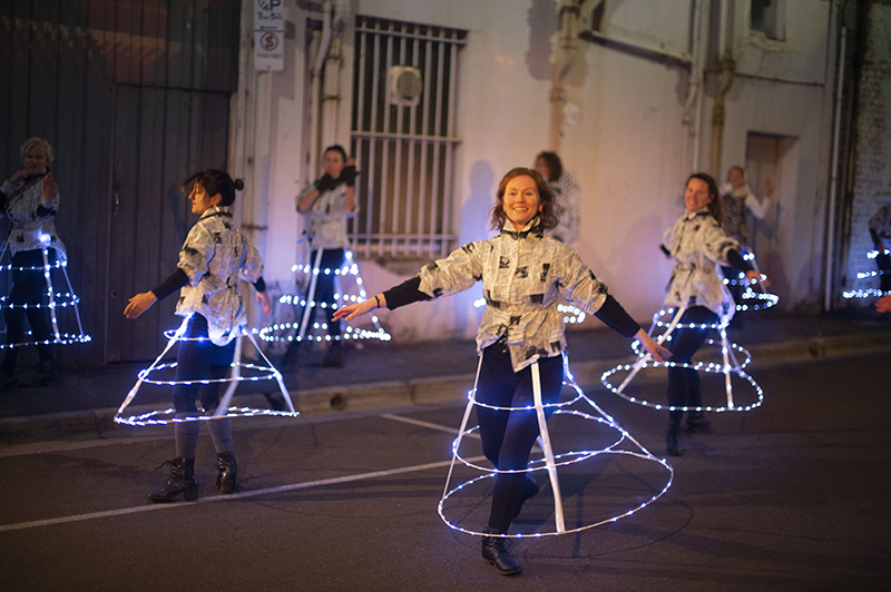 Several dancers dance on a dark city street with hoop skirts with LED lights.