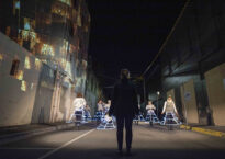 On a dark city street, a woman in black stands before several dancers wearing hoop skirts with LED lights.