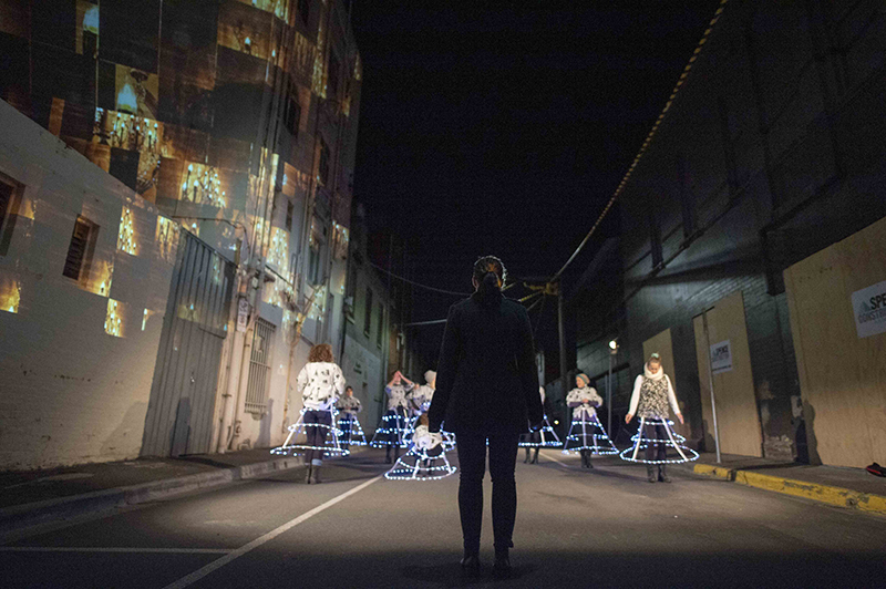 On a dark city street, a woman in black stands before several dancers wearing hoop skirts with LED lights.