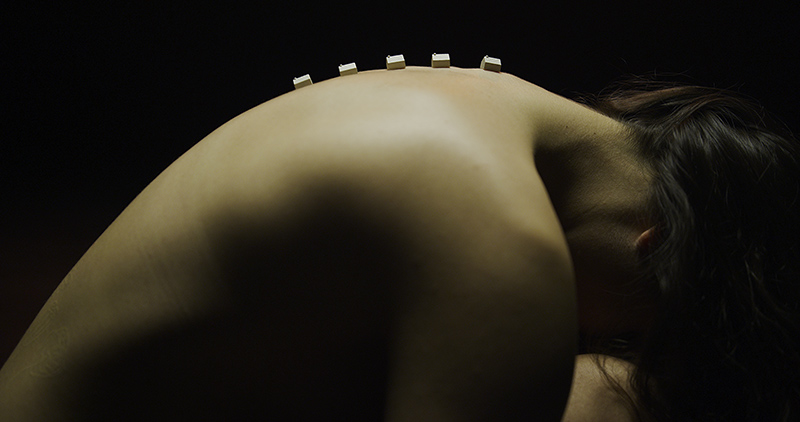 A naked back bathed in soft light from above with tiny houses on the curve of the upper spine.