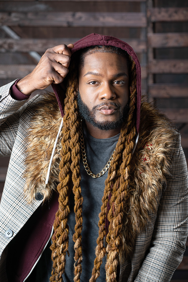 Malakhi is wearing a fur trimmed coat and long braids. He is slowly taking off a hood.