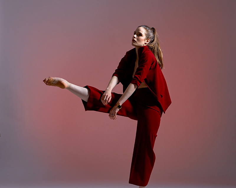 Megan wears a red suit and points her foot in front of her with her torso hunched over.