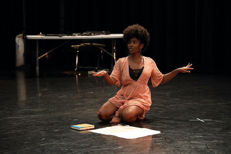 Melanie kneels onstage over papers and a book. She looks forward and her hands extend to the side.