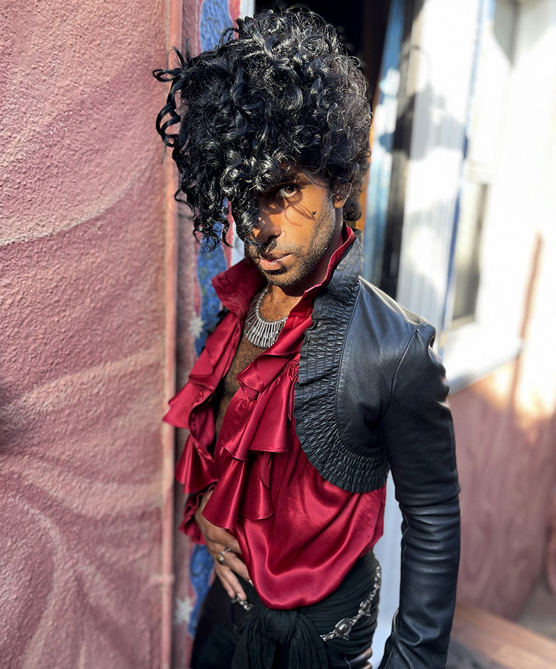 Nick close-up wearing a red frilly shirt and tight black jacket and pants with a whirl of curly hair in front of his face.