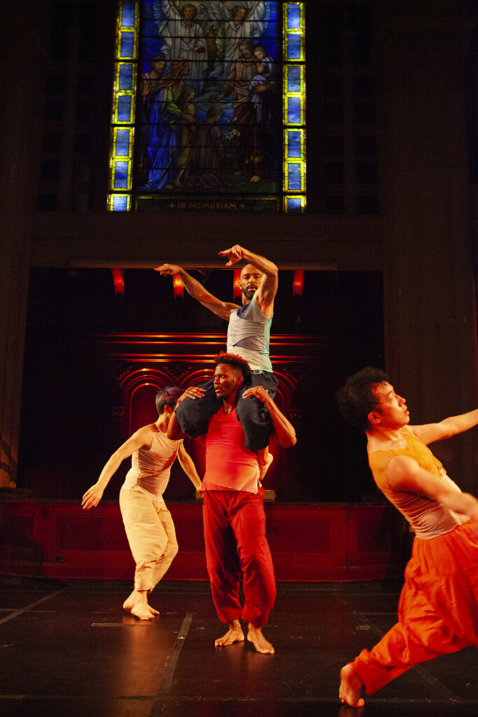 Four male dancers move in different directions in front of a blue stain glass window.
