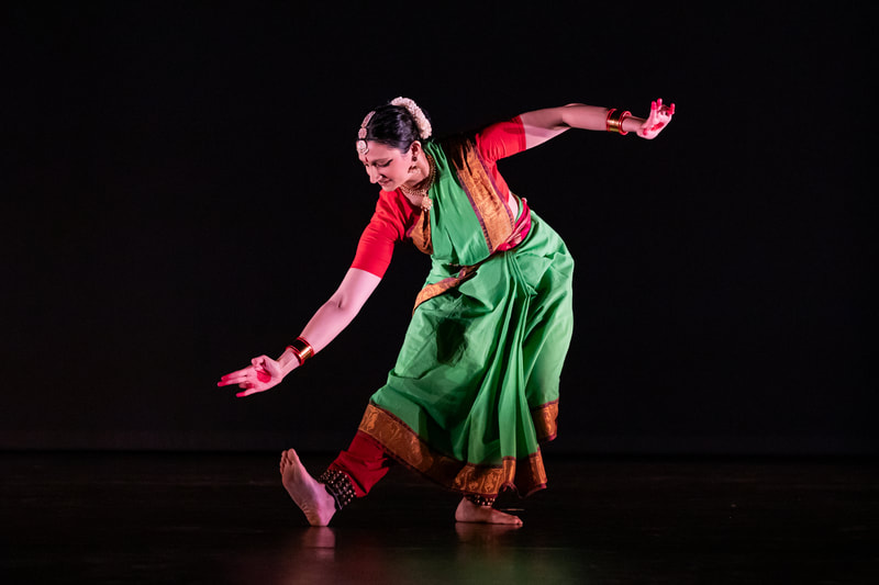 Sloka bends over one leg, gesturing toward her flexed foot. She wears a green and red sari.