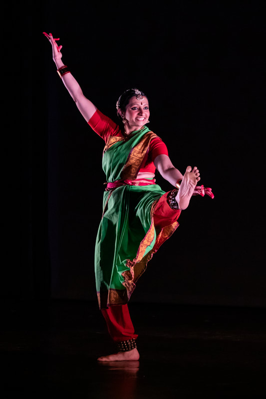 Sloka stands on one leg with the other flexed and extended out. Her hands open in a welcoming gesture. She is wearing a green and red sari.