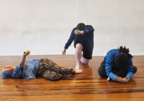 Three dancers are on a wood floor. One is laying down holding up a tennis ball. Another is kneeling and holding a tennis ball over their lowered head. A third is sitting cross legged and hunched over.