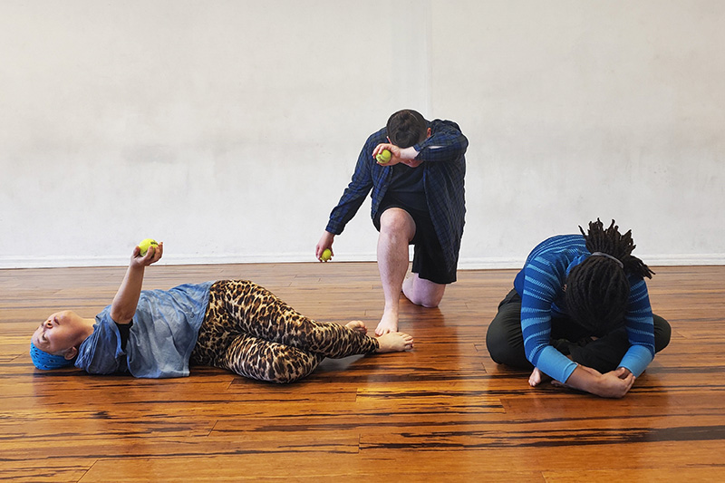 Three dancers are on a wood floor. One is laying down holding up a tennis ball. Another is kneeling and holding a tennis ball over their lowered head. A third is sitting cross legged and hunched over.