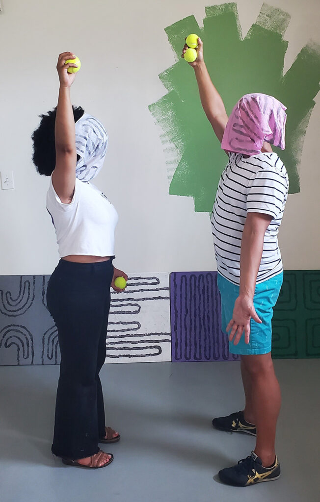 Two dancers stand and face each other. They are both masked and hold tennis balls above their heads.