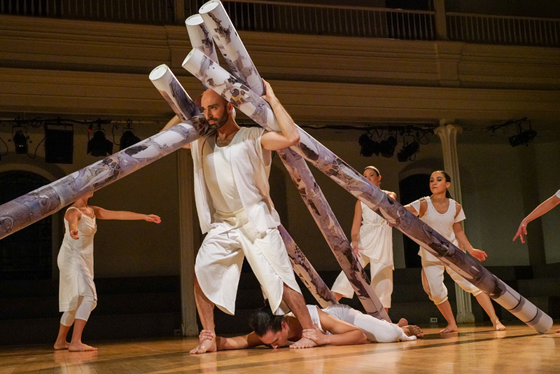 Dancers wearing white move around the stage lifting long tube-like structures.