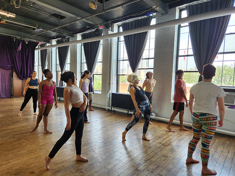8 students stand and follow the teacher in a dance studio with large dark curtains over bright windows.