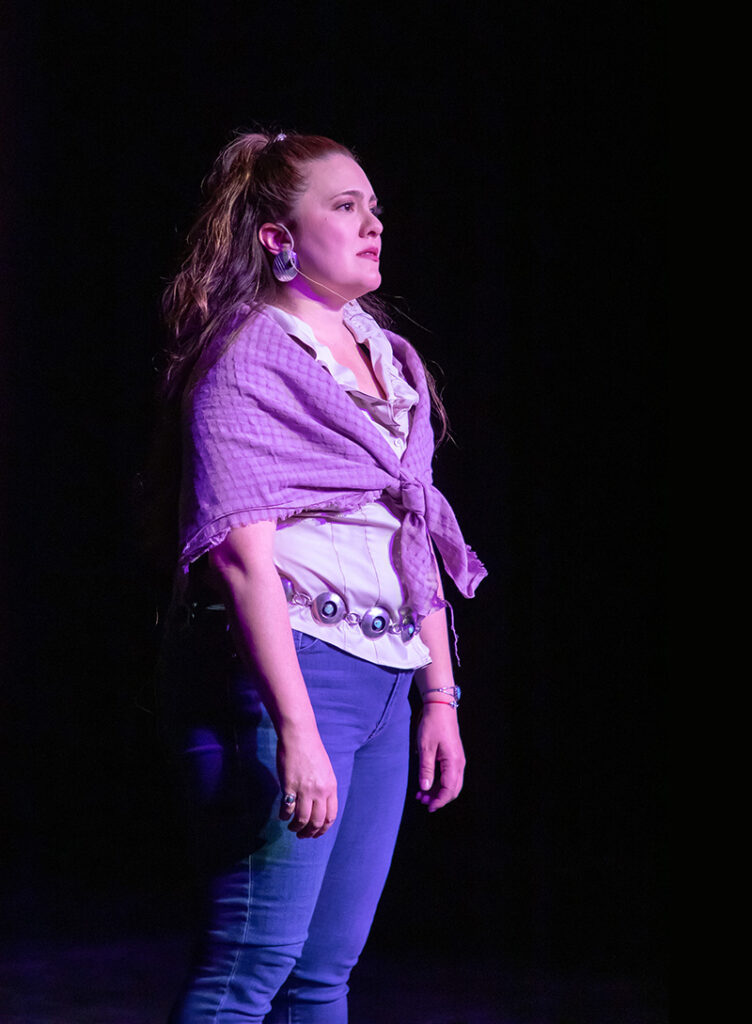 Yvonne stands and looks out from the stage. She wears jeans and a shawl.